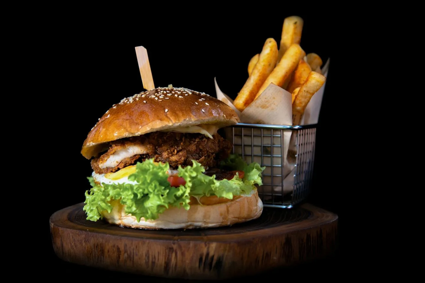 A tall burger with fries in a wire basket photographed against a black background