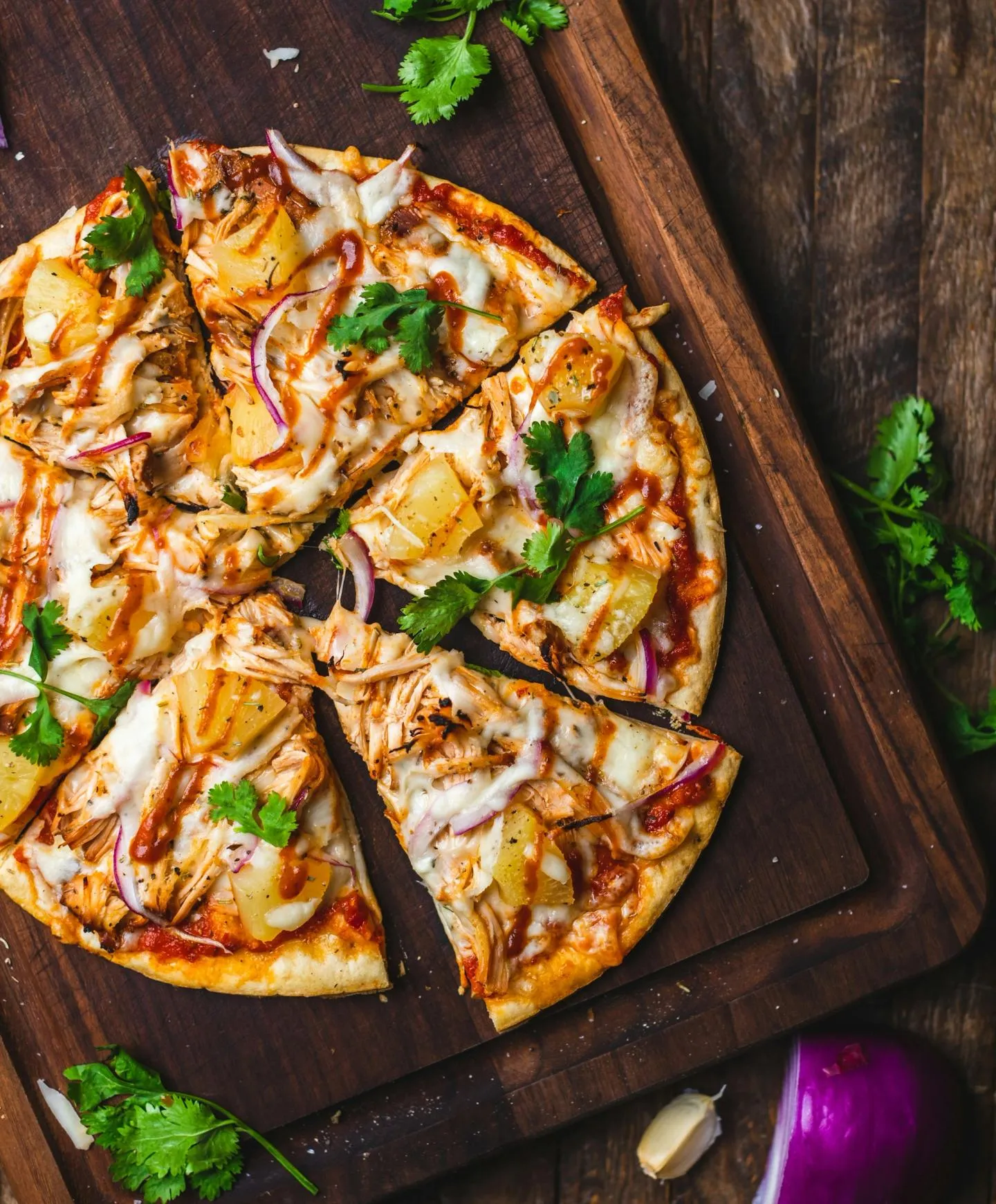 A thin-crust pizza sliced on a rustic wooden board with fresh herbs scattered around it