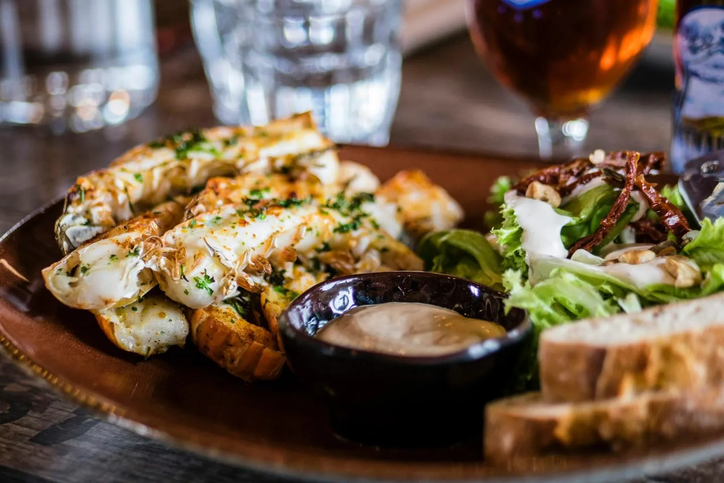 A wooden serving board with grilled fish, a bowl of dipping sauce, bread and a glass of beer