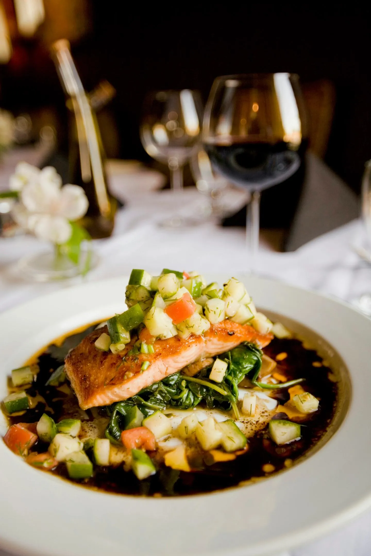 A seared salmon fillet on a bed of vegetables in a shallow white bowl, on a set restaurant table