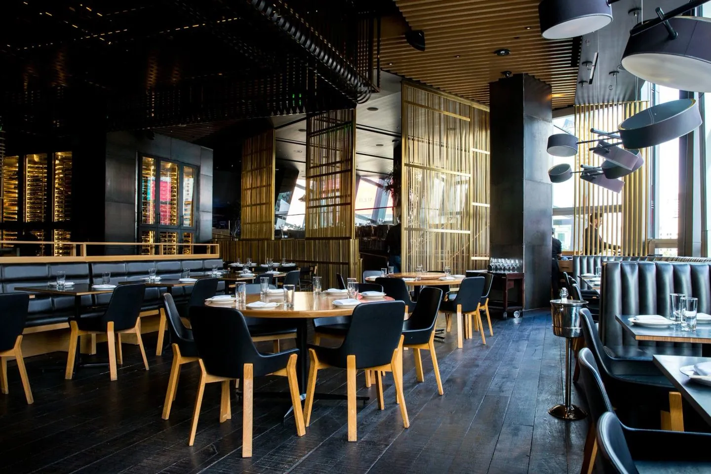 A modern restaurant dining room with warm wood tables, dark banquettes and pendant lighting