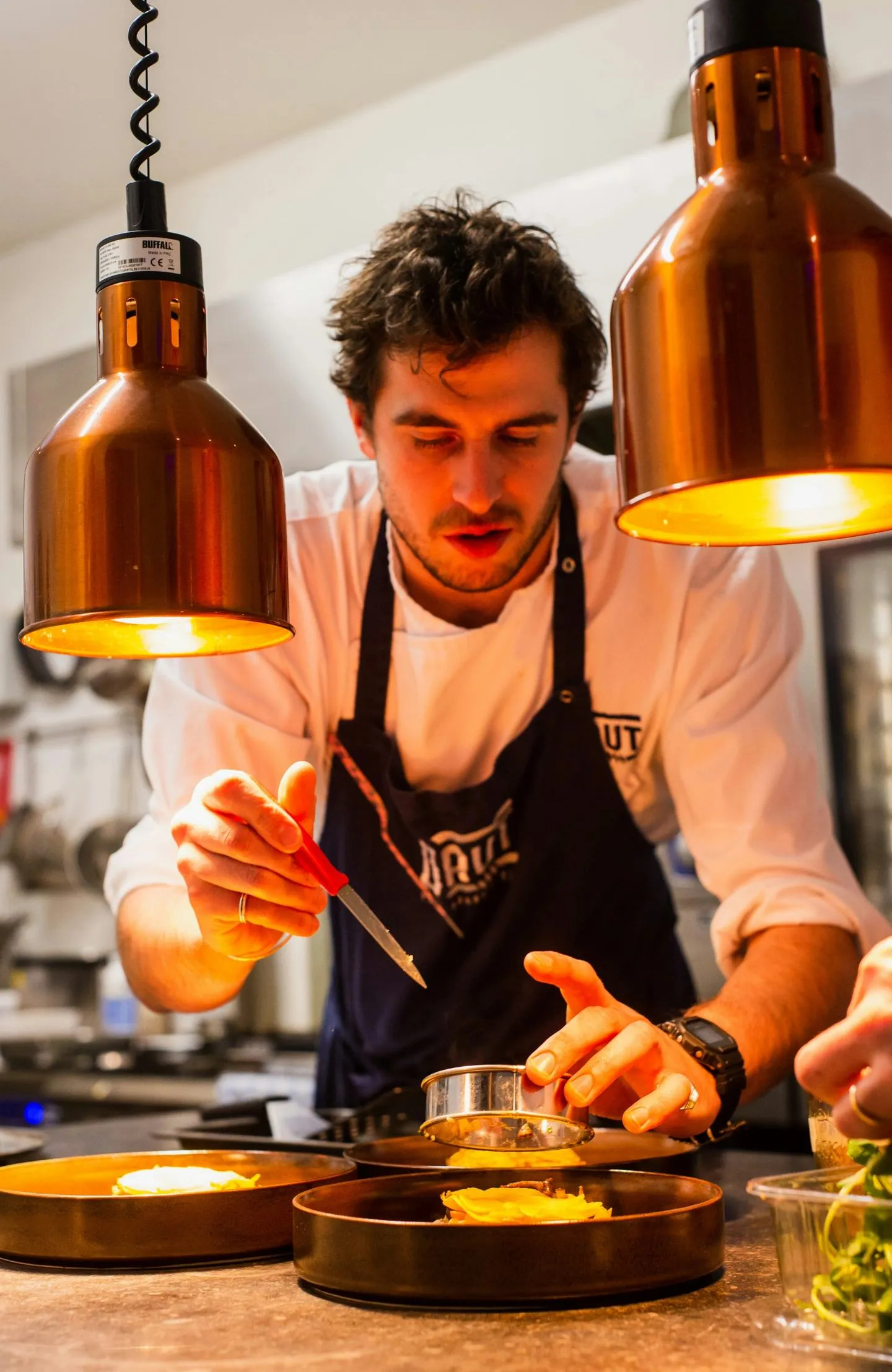 A cook in an apron plating a dish at a restaurant pass under warm copper pendant lamps
