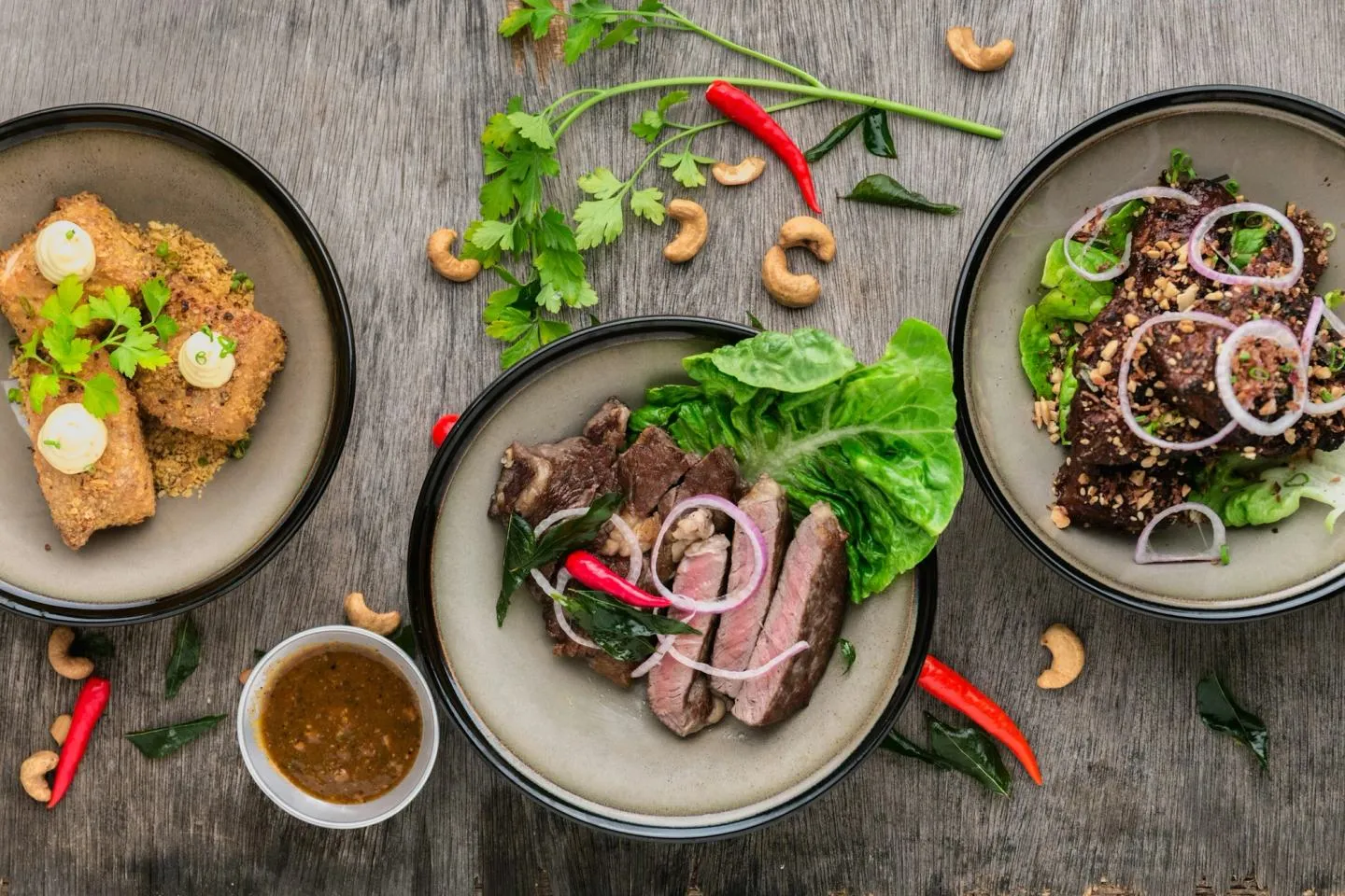 Overhead view of several grey ceramic plates with sliced meat, greens, cashews and a small bowl of sauce