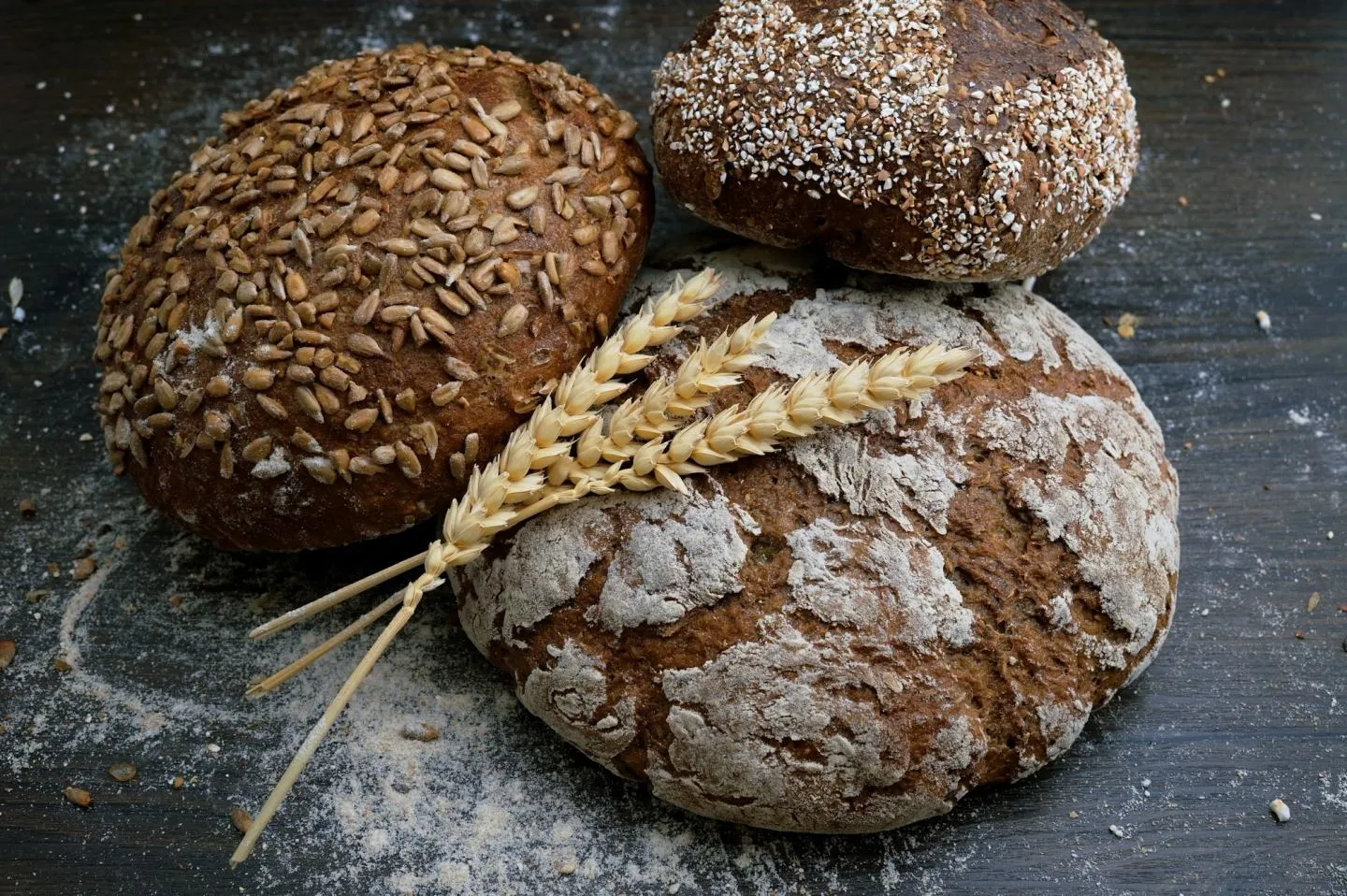 Three round sourdough loaves with scored crusts on a dark floured surface, with wheat stalks alongside