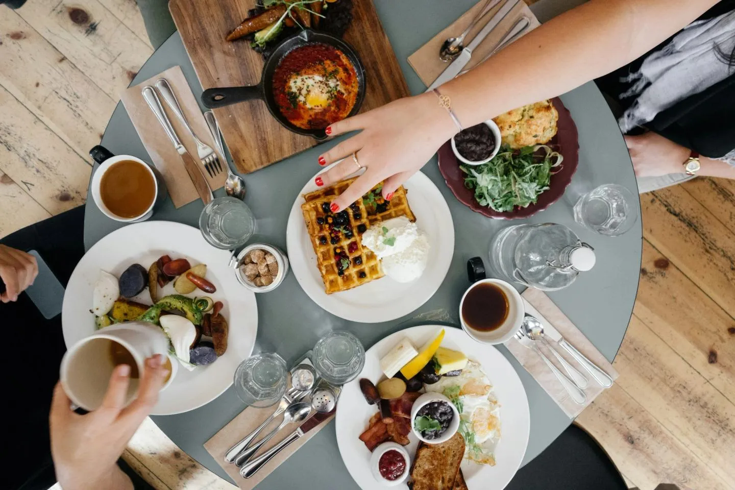 An overhead shot of a shared brunch table with waffles, coffee cups, small plates and several people reaching in